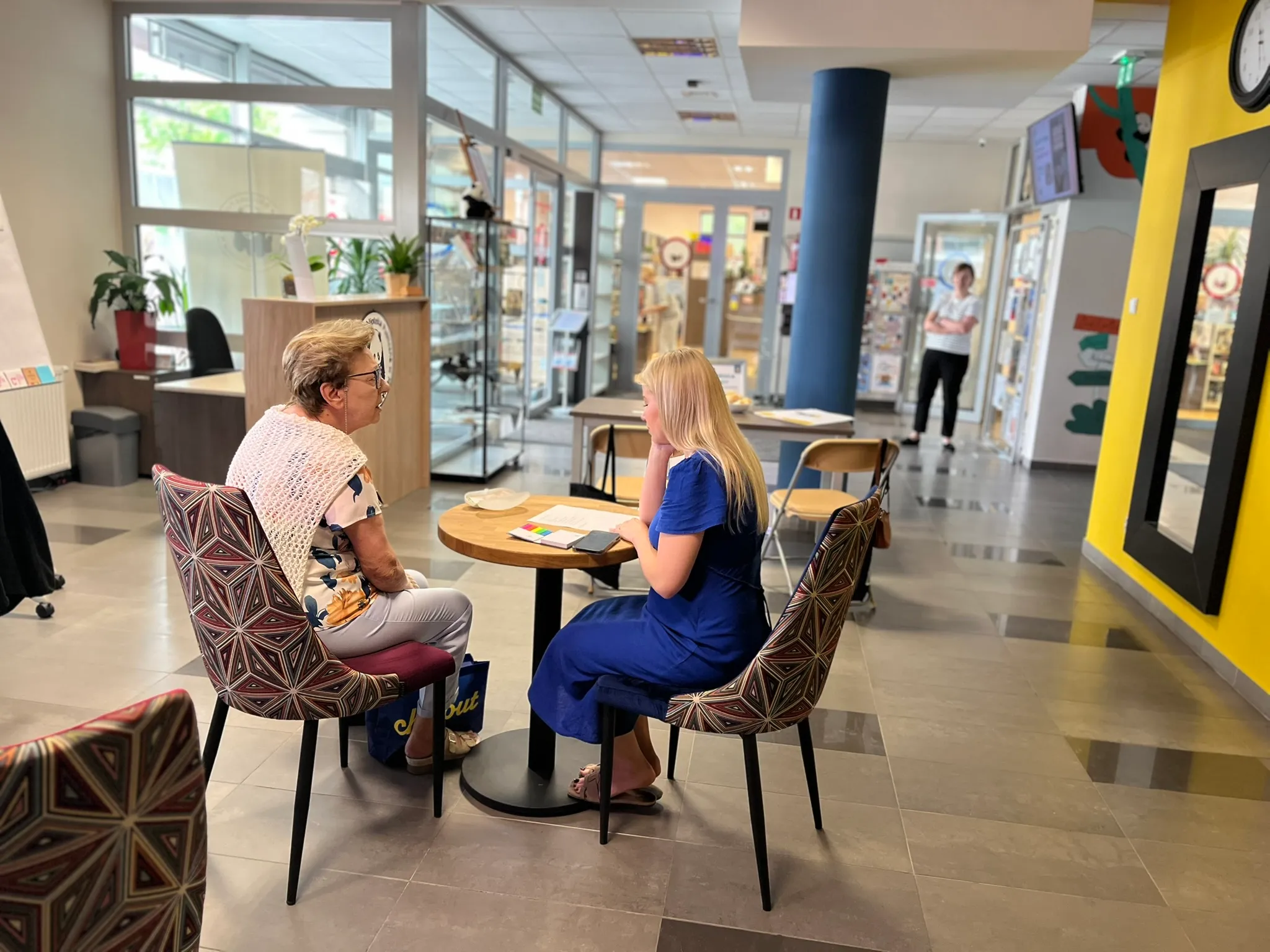 Dwie kobiety siedzą naprzeciwko siebie przy małym okrągłym stole w jasnym wnętrzu, w tle stoi trzecia osoba.