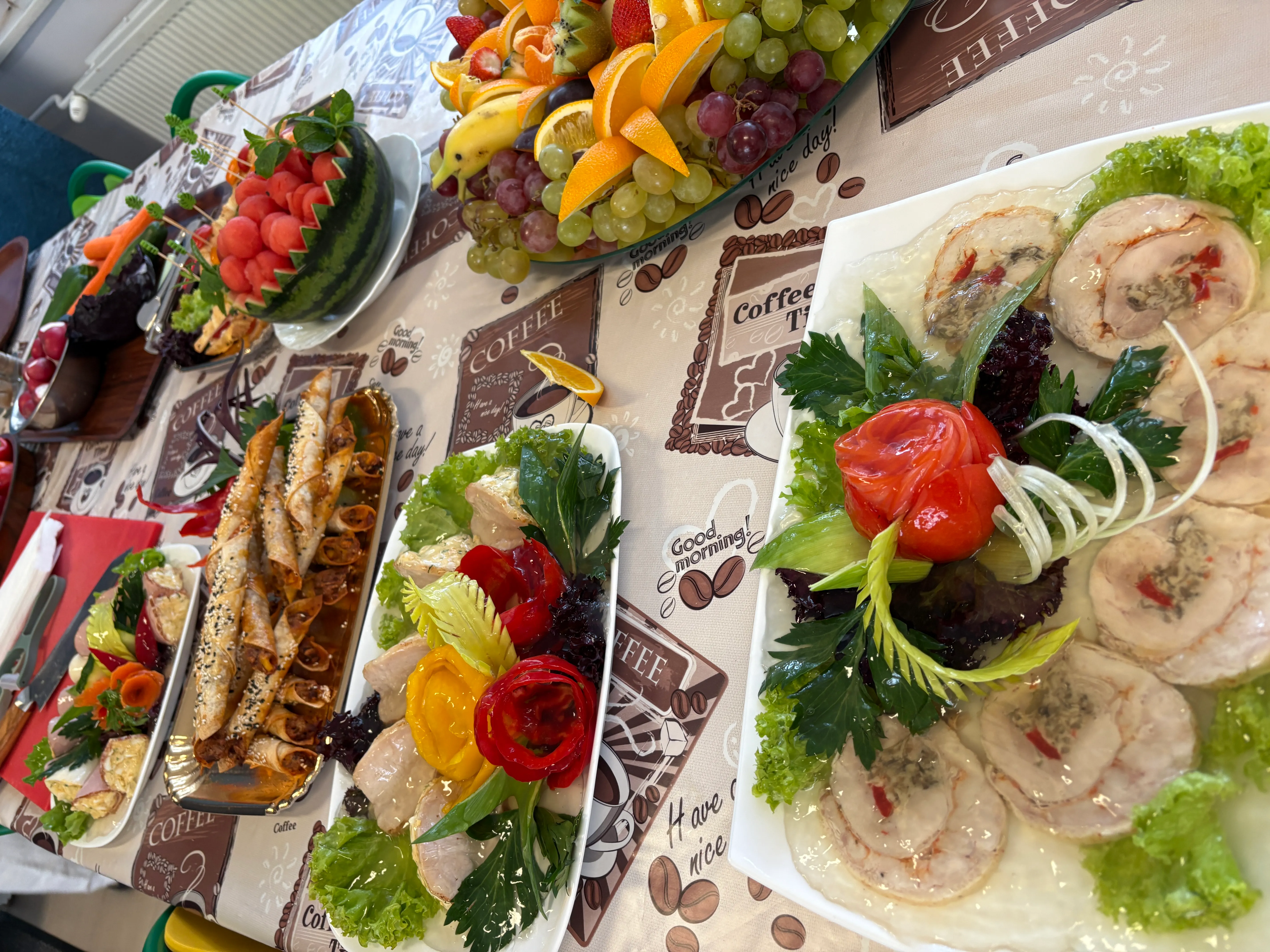 Stół nakryty obrusem z motywem kawy, na którym leżą talerze z różnorodnymi przekąskami i owocami. Po prawej plasterki roladek mięsnych z dekoracją z liści sałaty i warzywnych ozdób. W centrum różne przekąski na talerzach, między innymi roladki i sałatki z warzywami. Po lewej miska z wyciętymi kulkami arbuzów oraz talerz z kompozycją owoców takich jak winogrona, kiwi, pomarańcze i truskawki. Meble i wyposażenie w tle.