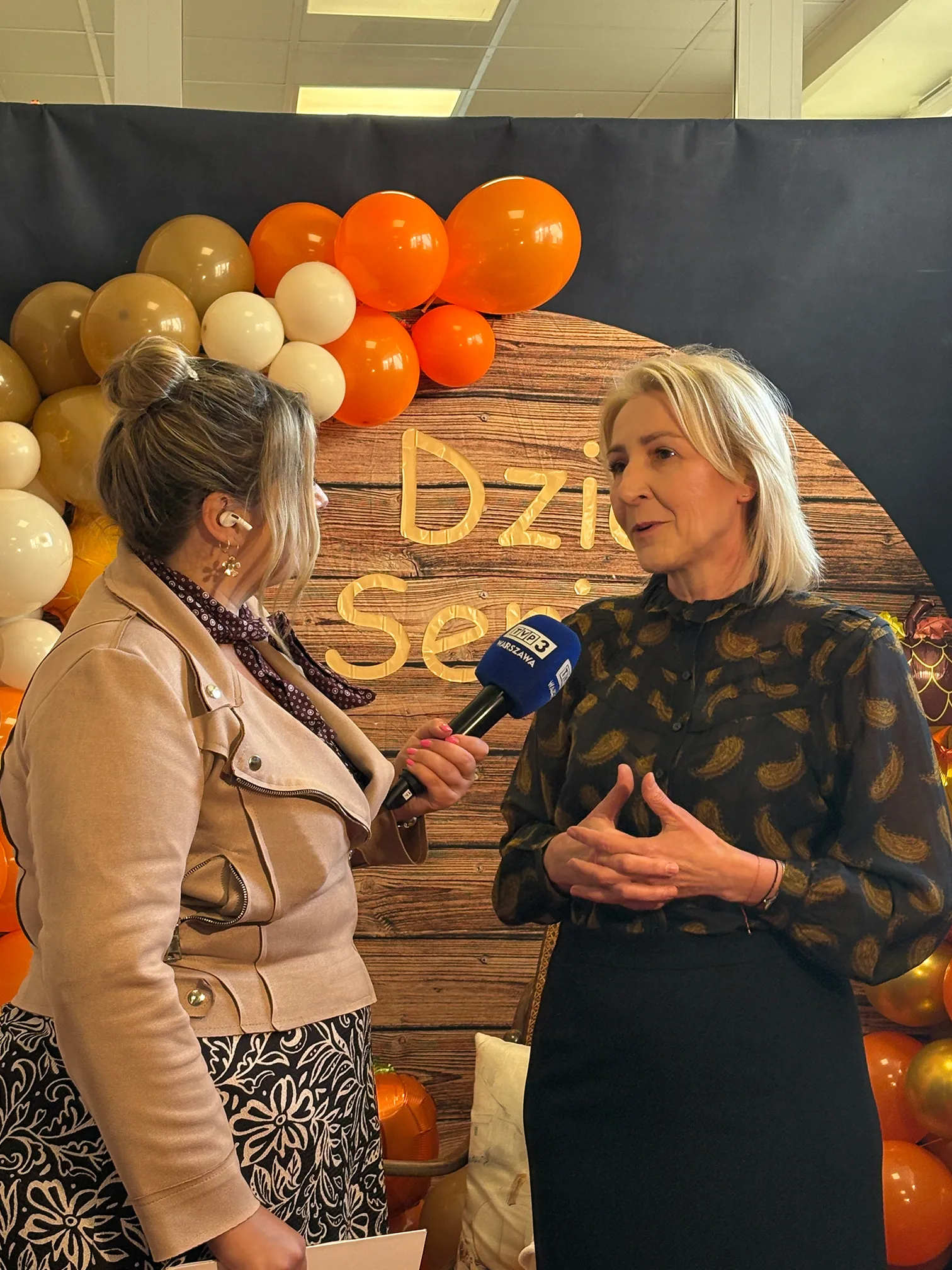 Dwie kobiety rozmawiają podczas wywiadu na tle drewnianej dekoracji i pomarańczowo-beżowych balonów. Jedna kobieta trzyma mikrofon z napisem TVP3 Warszawa. Druga kobieta gestykuluje podczas rozmowy. Scena odbywa się wewnątrz.