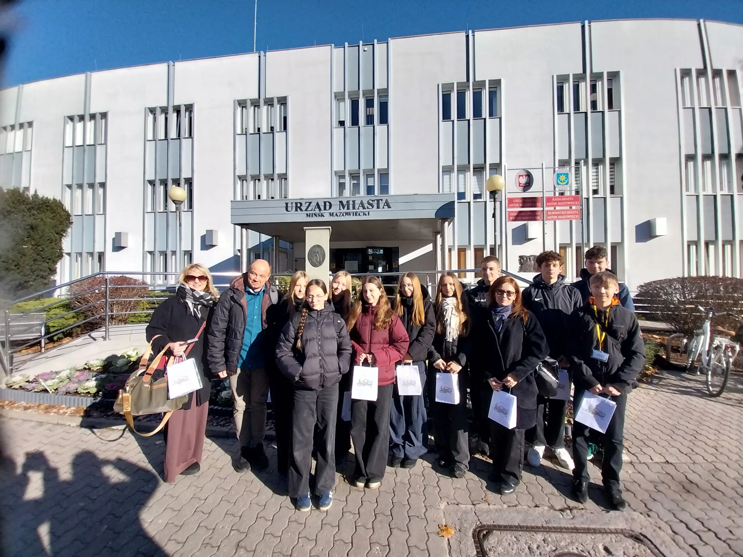 Grupa czternastu osób stoi przed białym budynkiem Urzędu Miasta Mińsk Mazowiecki w dzień, na chodniku z szarych kostek brukowych. Większość osób trzyma w rękach białe torby z nadrukiem. W tle widoczne dwa lampy uliczne i tablice informacyjne na ścianie budynku. Niebo jest bezchmurne, światło słoneczne oświetla całą scenę.