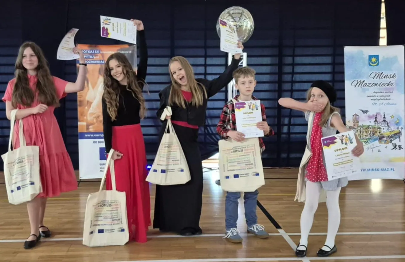 Pięcioro dzieci stoi na sali gimnastycznej. Trzy dziewczynki i chłopiec trzymają dyplomy i torby na ramionach. Jedna dziewczynka zasłania usta ręką. W tle widać dwa stojące banery i ciemne zasłony.