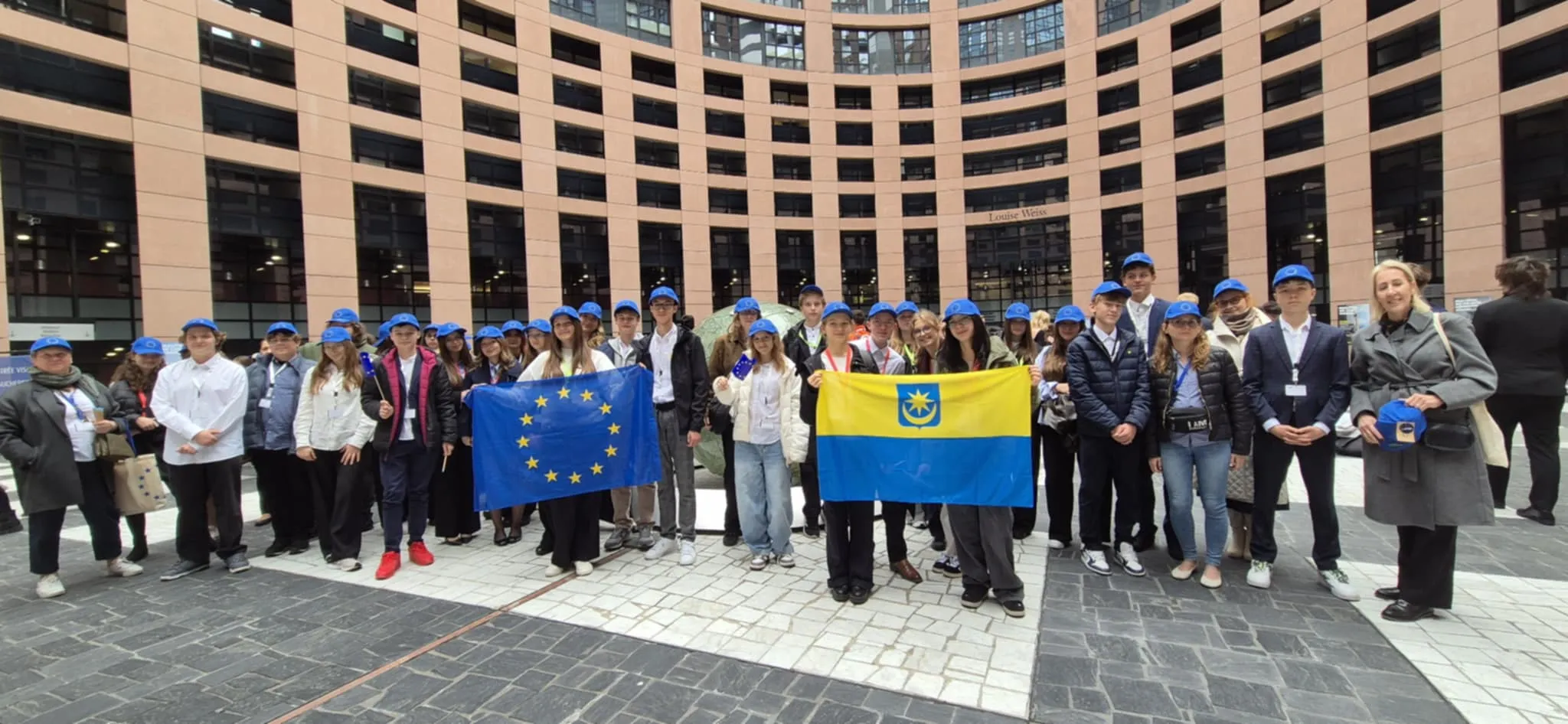 Grupa około 30 osób stojących na zewnątrz w półkolistym placu otoczonym przez nowoczesny budynek z dużymi oknami. Większość osób ma niebieskie czapki. Dwie osoby w centrum trzymają flagi – flagę Unii Europejskiej oraz flagę w żółto-niebieskich barwach z symbolem na żółtym polu. Podłoże z ciemnych i jasnych kamiennych płytek.