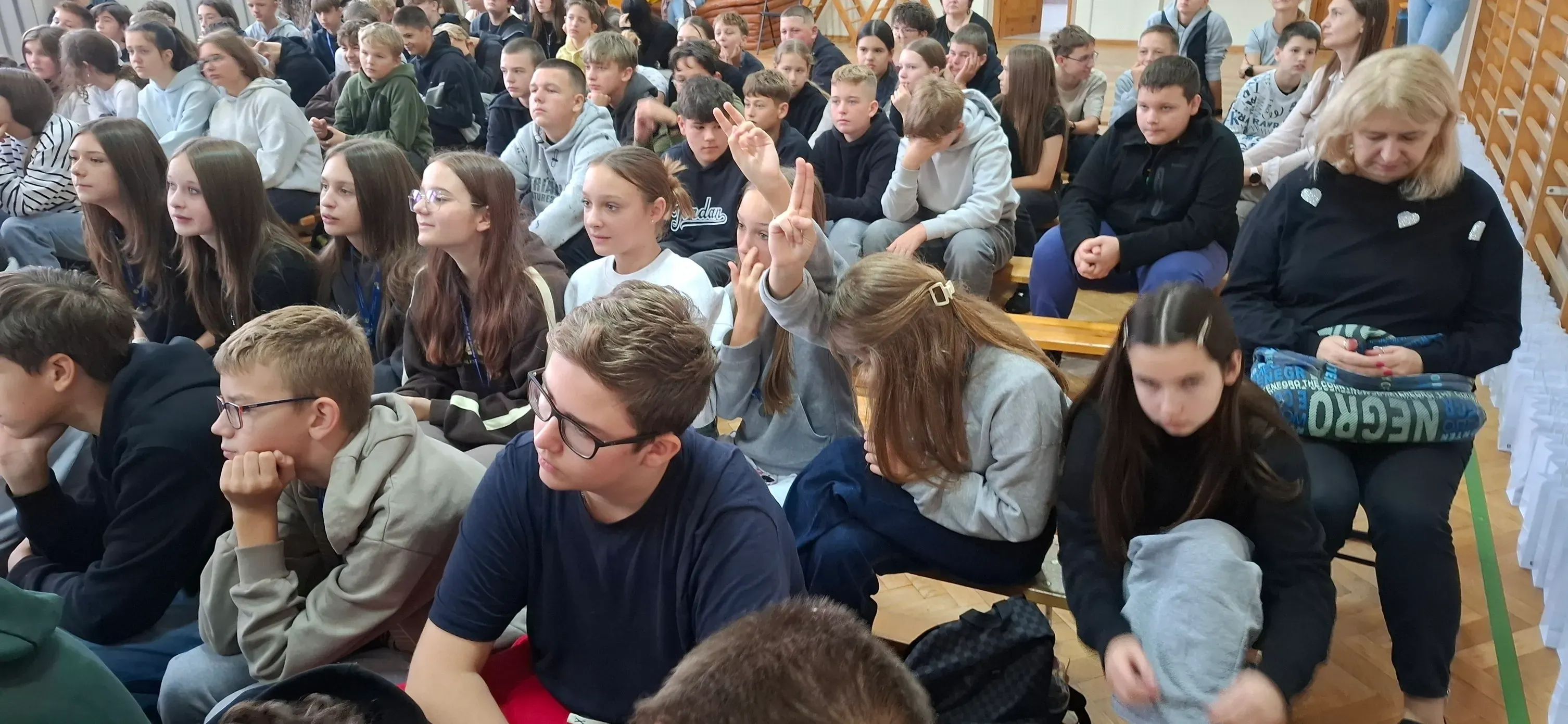 Duża grupa młodych osób siedzi na drewnianych ławkach w sali z drewnianymi panelami na ścianie, większość patrzy przed siebie, dwie dziewczyny wykonują znak zwycięstwa rękami, kobieta siedząca po prawej trzyma torbę na kolanach i patrzy w dół.