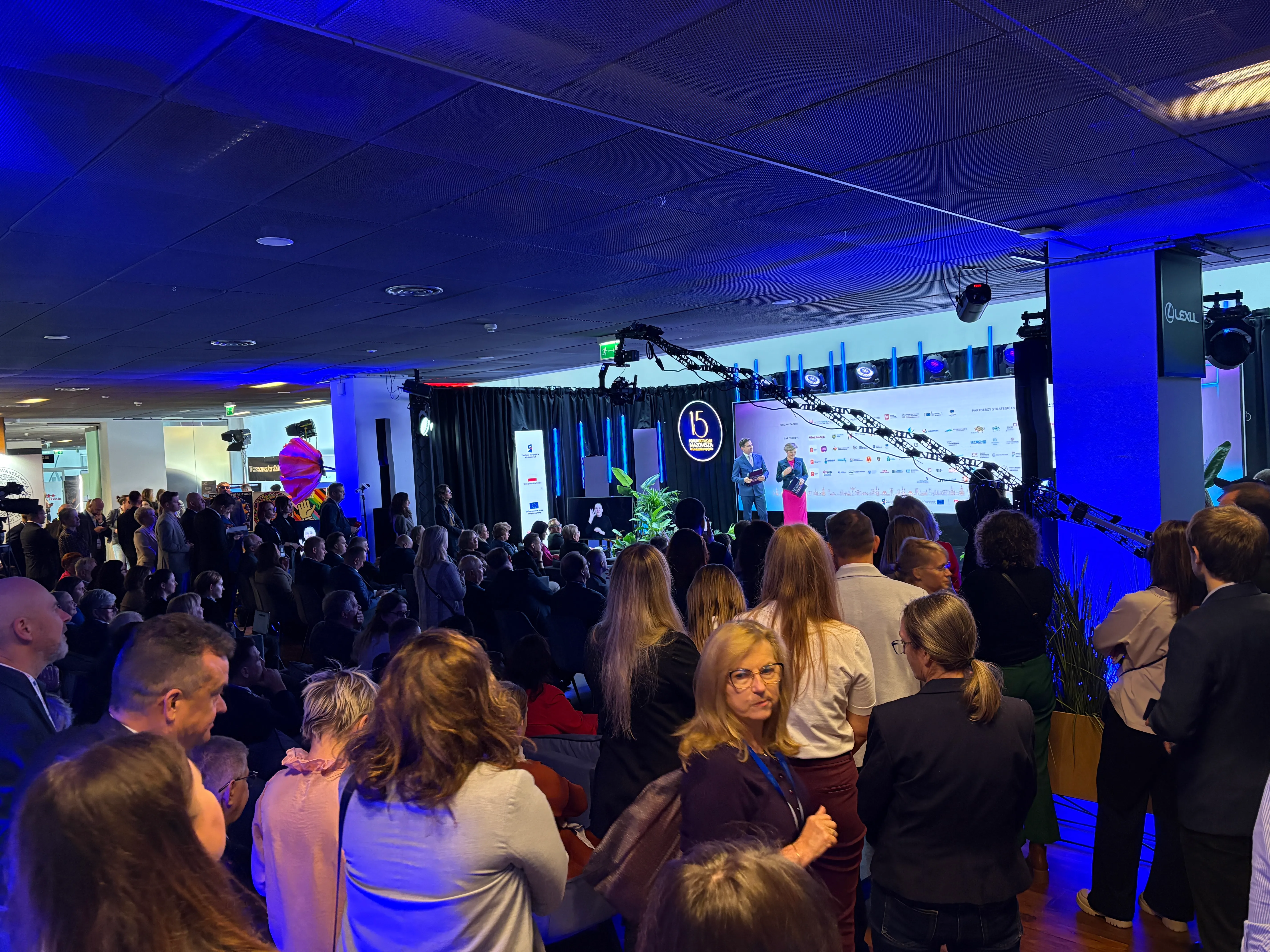 Duża grupa ludzi stoi i siedzi w hali konferencyjnej z niebieskim oświetleniem sufitowym, skupiona na dwóch prowadzących na scenie z logo "15" w tle.