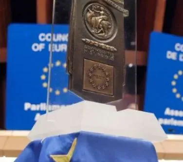 Medal z symbolem Unii Europejskiej zawieszony na przezroczystym stojaku, umieszczony na niebieskiej tkaninie z żółtą gwiazdą.