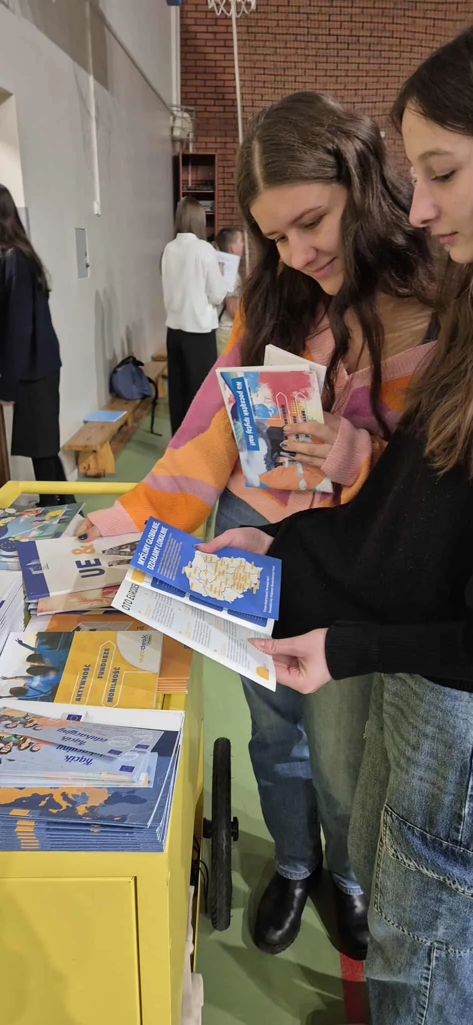 Dwie młode kobiety stoją przy żółtym stoisku i czytają broszury. Jedna ma pomarańczowo-różowy sweter, druga czarny sweter i jeansy. W tle widać inne osoby i czerwoną ścianę z cegły. Pomieszczenie jest jasno oświetlone.