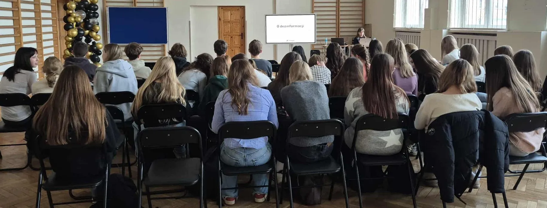 Grupa około trzydziestu osób siedzi na krzesłach w szkolnej sali gimnastycznej, zwrócona przodem do nauczycielki i ekranu z napisem „O dezinformacji”. Pomieszczenie jest jasne, z oknami po prawej stronie i drewnianą ścianą z drabinkami po lewej. Po lewej stronie stoi dekoracja z czarno-złotych balonów.