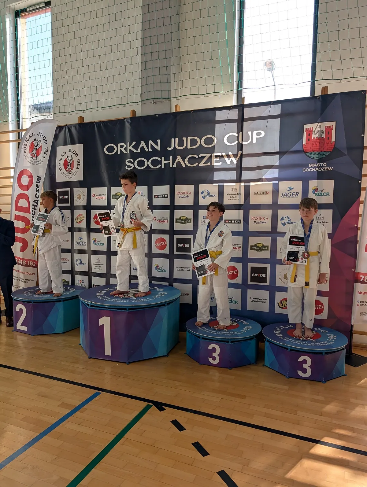 Czterech chłopców w białych judogach z żółtymi pasami stoi na podium w hali sportowej. Za nimi jest czarne tło z napisem „ORKAN JUDO CUP SOCHACZEW” i logo miasta Sochaczew. Chłopiec na najwyższym podium trzyma dyplom i medal, pozostali także mają medale i dyplomy. Podium jest oznaczone numerami 1, 2 i dwukrotnie 3.