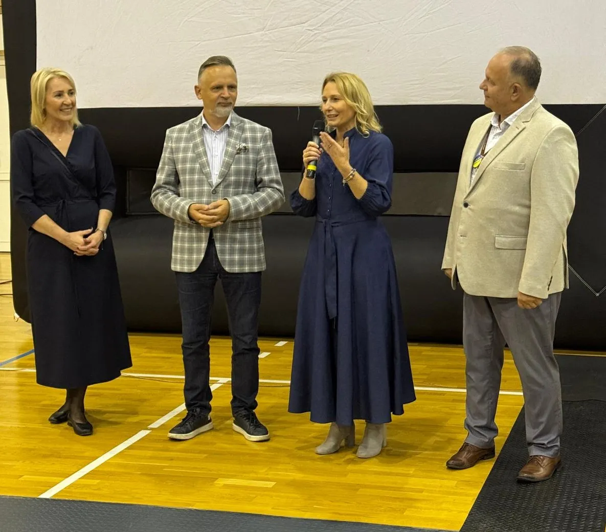 Cztery osoby stoją na drewnianej podłodze w sali, trzecia od lewej mówi do mikrofonu, pozostałe trzy słuchają. Wszystkie osoby są ubrane formalnie. Tło jest czarno-białe.