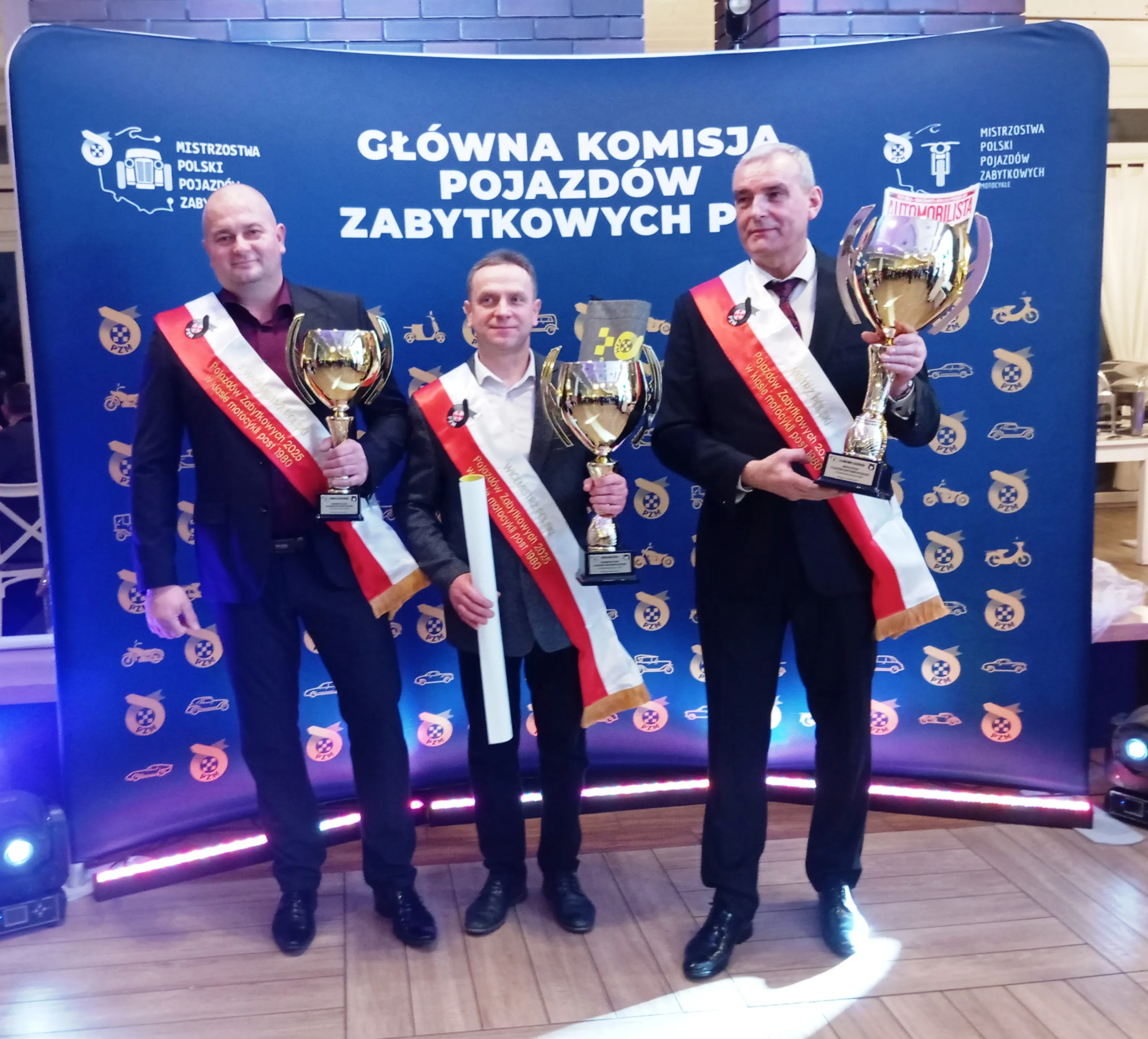 Trzech mężczyzn stoi na tle niebieskiego banera z napisem i ikonami pojazdów. Każdy z nich ma na sobie czarne ubranie z biało-czerwonymi szarfami i trzyma duży złoty puchar. Wnętrze pomieszczenia oświetlone jest sztucznym światłem.