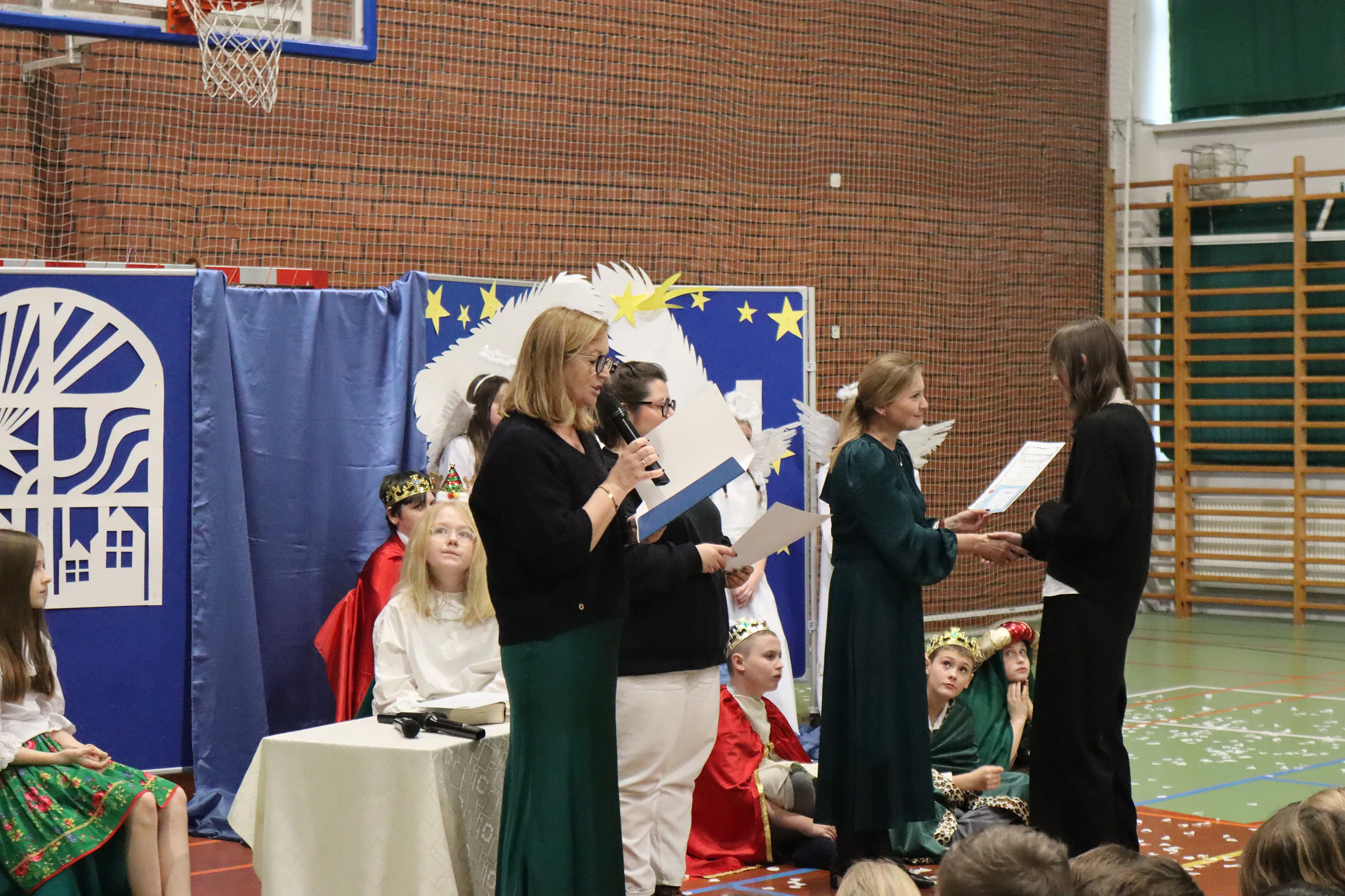 Kilka osób uczestniczy w uroczystości w sali gimnastycznej, jedna kobieta czyta z mikrofonem, inna wręcza dokument młodej osobie. W tle dzieci w kostiumach aniołów i króli siedzą na podłodze. Na ścianie dekoracja z gwiazdami i sylwetką białego konia.