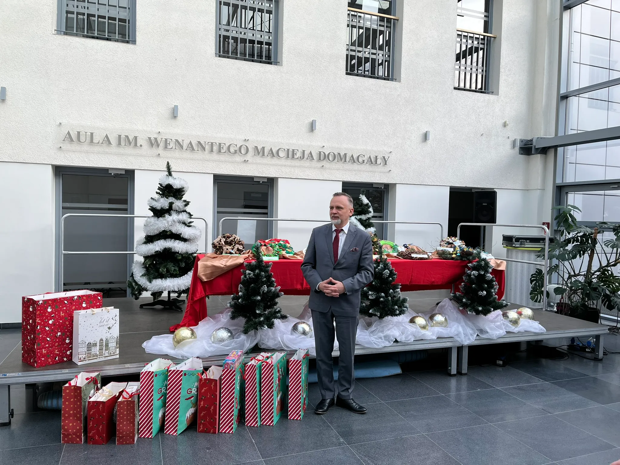 Mężczyzna w garniturze stoi obok sceny udekorowanej sztucznymi choinkami, białą tkaniną i dużymi złotymi oraz srebrnymi bombkami. Na scenie znajduje się stół z czerwonym obrusem i dekoracjami. Przed sceną stoi kilka kolorowych toreb prezentowych. W tle na ścianie widoczny jest napis "AULA IM. WENANTEGO MACIEJA DOMAGAŁY". Wnętrze jest jasne, z dużymi oknami po prawej stronie.