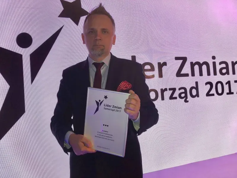 Mężczyzna w garniturze z bordową poszetką trzyma w dłoniach ramkę z dyplomem „Lider Zmian Samorząd 2017”.
