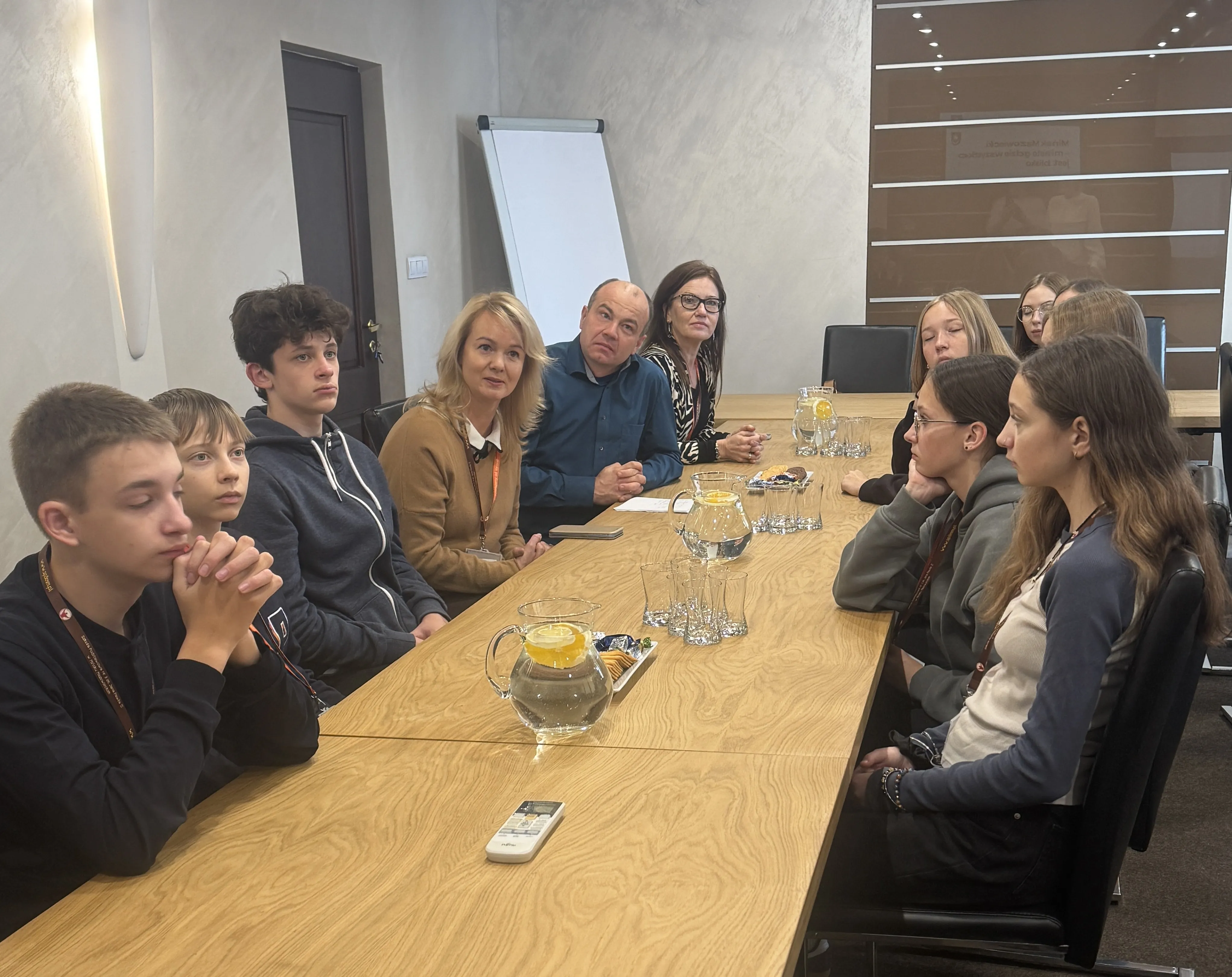 Dziesięć osób siedzi przy długim drewnianym stole w sali konferencyjnej. Na stole znajdują się szklanki, dzbanki z wodą i cytryną oraz przekąski. W tle stoi flipchart. Ściany mają jasny kolor, a w pokoju jest sztuczne oświetlenie.