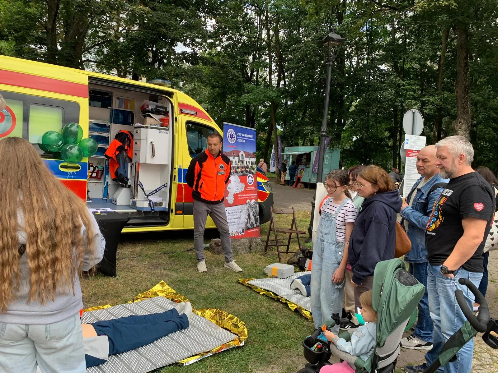 Grupa osób obserwuje pokaz pierwszej pomocy na trawie obok otwartej karetki w parku podczas dnia.
