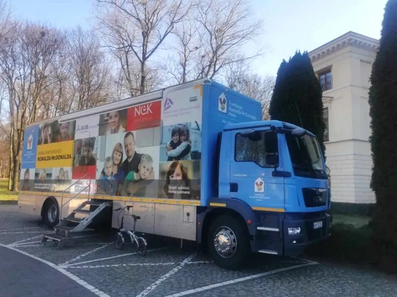 Niebieski samochód ciężarowy z przyczepą oklejoną zdjęciami rodzin i logo Fundacji Ronalda McDonalda zaparkowany przy chodniku, obok schodów i roweru.