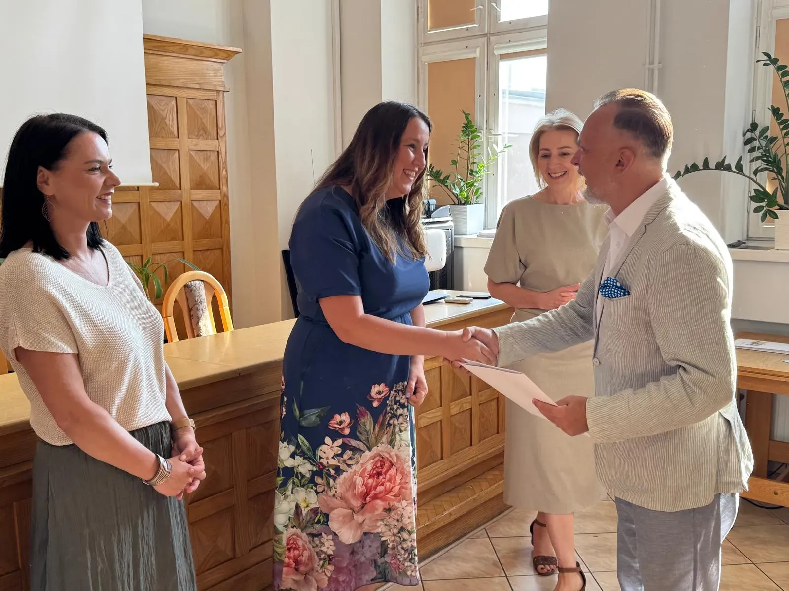 Cztery osoby stoją w jasnym pomieszczeniu. Mężczyzna w jasnej marynarce podaje dokument kobiecie w sukience w kwiaty. Obok stoją dwie uśmiechnięte kobiety.