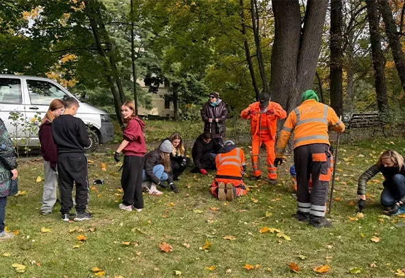 Grupa osób pracuje na trawniku w parku wśród drzew, część osób ubrana jest w pomarańczowe kamizelki i kombinezony robocze, inne w ubrania codzienne. Niebo jest częściowo zakryte przez liście drzew, dzień jest pochmurny.