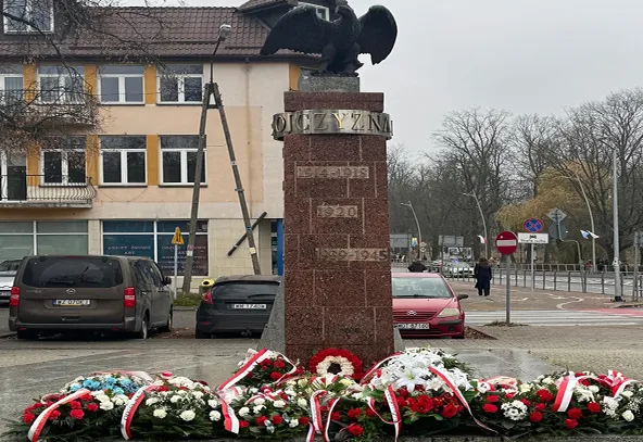 Pomnik orła stojący na ceglanym cokole na placu z wieńce kwiatów u jego podstawy, w tle budynki i zaparkowane samochody.