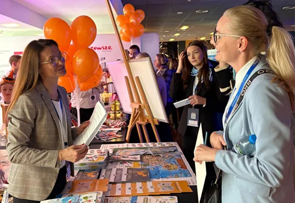 Dwie kobiety rozmawiają przy stoisku z ulotkami i plakatami wewnątrz budynku. W tle inne osoby, pomarańczowe balony i tablica na sztaludze.