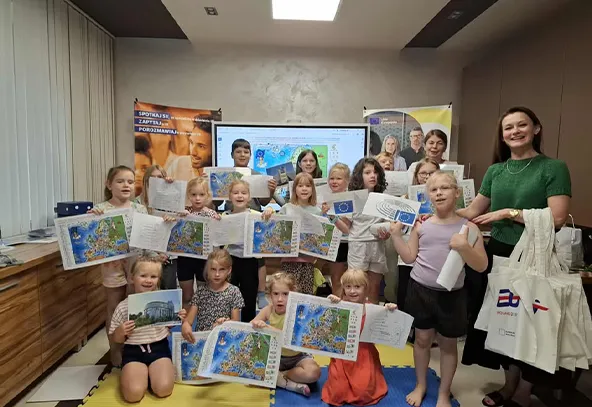 Grupa dzieci i dwóch dorosłych trzymających mapy i broszury, stojących i siedzących na żółtej macie w pomieszczeniu.