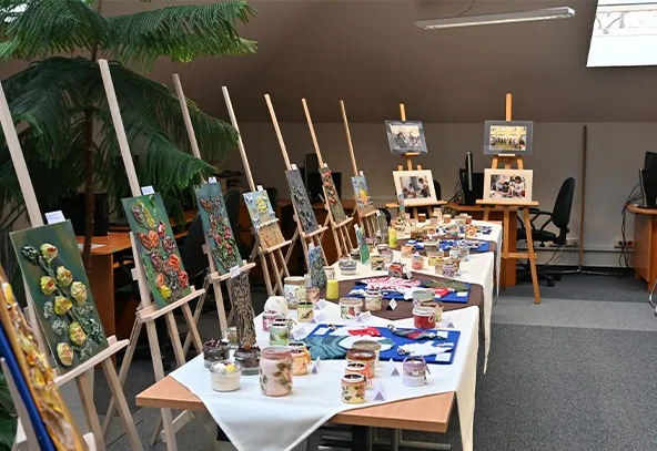 Wnętrze z białym stołem nakrytym obrusami. Na stole ustawione słoiki z ozdobami oraz niebieskie podkładki z ozdobami. Po lewej stronie stołu znajduje się sześć sztalug z kolorowymi obrazami kwiatów. W głębi dwa większe obrazy na dwóch sztalugach. W tle biurka, krzesła i roślina doniczkowa. Pomieszczenie oświetlone światłem dziennym przez okno dachowe.