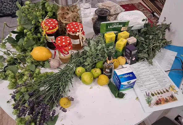 Na okrągłym stole znajduje się kilka pęków suszonych ziół, trzy sloiki z czerwonymi pokrywkami, cytrusy, tabletki i butelka z kroplomierzem oraz dokument z wykazem roślin.