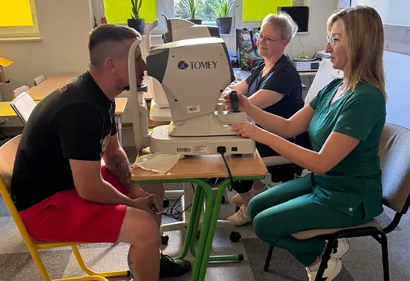 Mężczyzna przechodzi badanie wzroku przy użyciu urządzenia medycznego, obsługiwanego przez kobietę w zielonym uniformie, podczas gdy inna kobieta obserwuje badanie przy stole w dobrze oświetlonym pomieszczeniu.