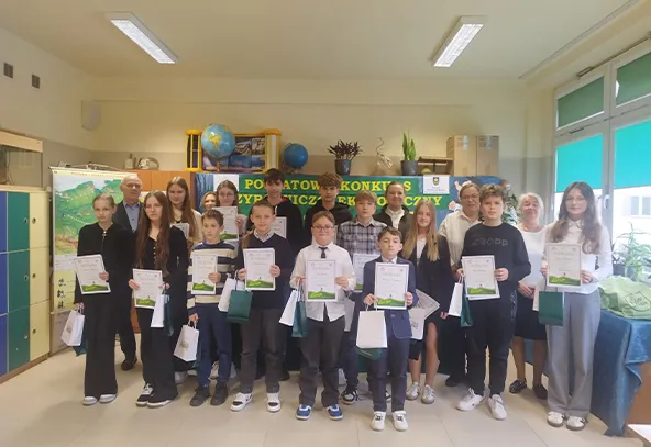 Grupa 17 osób w pomieszczeniu o jasnych ścianach stoi w dwóch rzędach, trzyma dyplomy i torby. W tle zielony plakat z żółtym napisem. W pomieszczeniu widoczne są półki z globusami i rośliny.