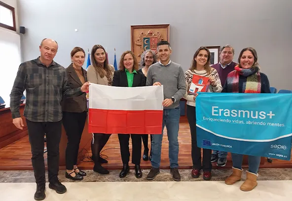 Grupa dziewięciu osób stoi wewnątrz jasnego pomieszczenia z drewnianą podłogą i szarymi ścianami. Dwie osoby trzymają z przodu biało-czerwoną flagę Polski, a dwie inne niebieski banner z napisem Erasmus+. W tle widoczna jest duża ramka z herbem oraz dwie flagi stojące przy ścianie.