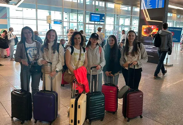 Sześć kobiet stoi w hali lotniska z walizkami na kółkach. W tle widać tablice informacyjne, dużą szybę i innych pasażerów. Oświetlenie dzienne, wnętrze jasne.