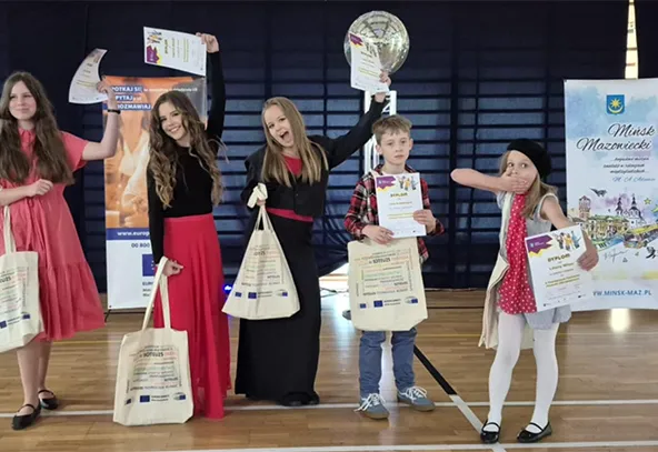 Pięcioro dzieci stoi w hali sportowej na parkiecie. Trzy dziewczynki z lewej strony trzymają torby i dyplomy, jedna z nich unosi dyplom wysoko. Chłopiec i dziewczynka po prawej stronie również trzymają dyplomy i torby. Dziewczynka po prawej robi gest zasłaniania ust ręką. W tle widoczne są dwa pionowe banery.