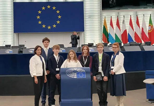 Siedem osób stoi przed mównicą w sali konferencyjnej z dużą flagą Unii Europejskiej na ścianie i zestawem flag państw członkowskich w tle. Wśród nich są młodzi ludzie oraz dwie dorosłe kobiety. Na mównicy widoczny jest symbol Parlamentu Europejskiego. W tle stoi osoba z aparatem fotograficznym.