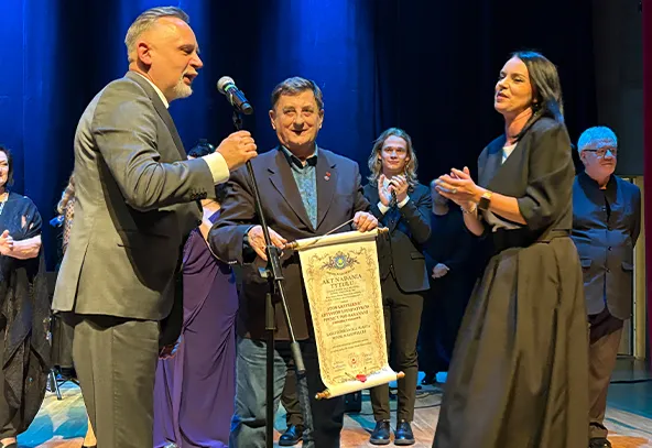 Na scenie teatralnej stoi siedem osób. Trzech mężczyzn i kobieta stoją na pierwszym planie. Jeden mężczyzna trzyma mikrofon, drugi mężczyzna trzyma zwinięty dokument w rękach. Kobieta z prawej strony klaszcze. W tle widać inne osoby, również stojące na scenie, a tło jest ciemnoniebieskie.