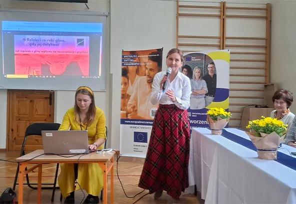 Kobieta w żółtej sukience siedzi przy stole i obsługuje laptopa. Obok stoi kobieta w białej bluzce i czerwonej spódnicy w kratę, trzyma mikrofon i przemawia. W tle dwa banery informacyjne. Po prawej stronie długi stół z białym obrusem i żółtymi kwiatami. Sala gimnastyczna z drabinką na ścianie.