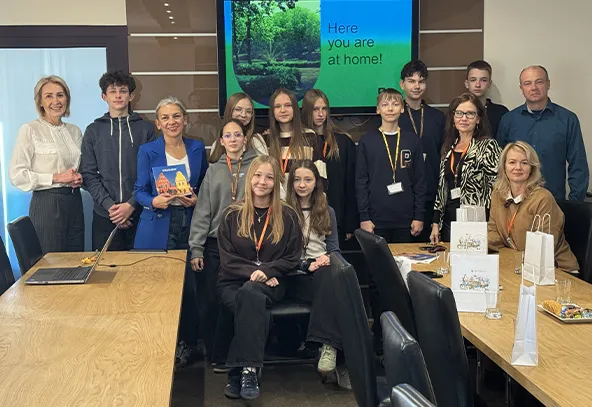 Grupa piętnastu osób w sali konferencyjnej. Dwanaście osób stoi na tle ściany z dużym ekranem pokazującym zieleń i napis „Here you are at home!”, trzy siedzą na krzesłach przy stole. W pomieszczeniu jasne oświetlenie, na stole torby papierowe i laptop.