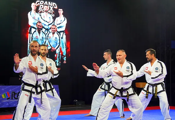 Pięciu mężczyzn w białych kimono z czarnymi pasami ćwiczy sztuki walki na niebiesko-czerwonej macie. Za nimi znajduje się duży, pionowy plakat z czarnym tłem i zdjęciem kilku osób w podobnych strojach oraz napisem „Grand Prix Polski”. Tło sceny jest ciemne.
