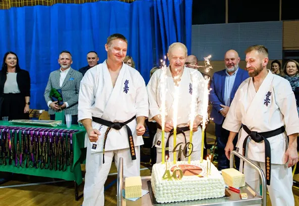 Trzech mężczyzn w białych kimonach z czarnymi pasami stoi wokół dużego tortu z dekoracjami i zapalonymi świecami iskrzącymi. W tle stoi grupa osób obserwujących wydarzenie w hali z niebieską zasłoną. Na stole leżą medale.