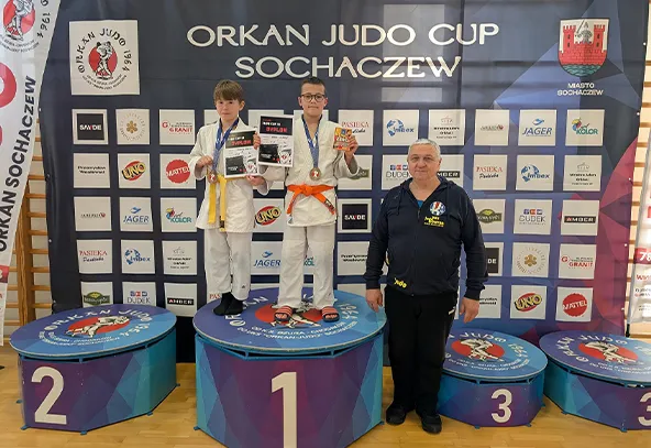 Dwoje dzieci w judogach stoi na podium z numerami 1 i 2, trzyma medale i dyplomy. Obok na podium stoi mężczyzna w granatowej bluzie. W tle baner z napisem Orkan Judo Cup Sochaczew oraz logotypy sponsorów.