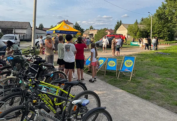 Grupa ludzi i dzieci stoi na zewnątrz obok rzędu zaparkowanych rowerów, w tle namiot i stoisko na trawniku przy słonecznej pogodzie.