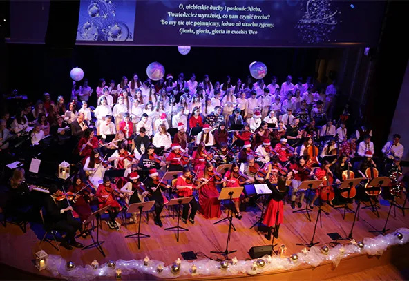 Na scenie znajduje się duża grupa muzyków i chórzystów. Muzycy grają na skrzypcach, wiolonczelach oraz innych instrumentach smyczkowych, większość ubrana jest na czerwono. Chórzystów jest wielu, stoją za muzykami, ubrani na biało. Scena jest podświetlona fioletowym światłem, a na dole znajduje się dekoracja z lampionami i światełkami. W tle na ekranie wyświetlony jest tekst.