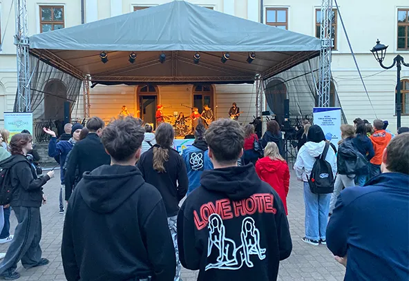 Grupa osób stoi na placu przed sceną z zadaszeniem, na której gra zespół muzyczny. Scena znajduje się przed jasnym budynkiem z oknami, jest wieczór. Widzowie mają na sobie różne ubrania, w tym czarne bluzy i kurtki.