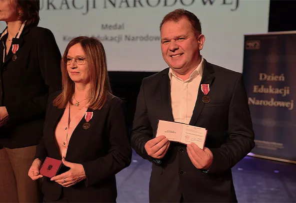 Dwóch uśmiechniętych dorosłych osób stoi na scenie w formalnych ubraniach, oboje mają przypięte medale na piersiach i trzymają dokumenty. W tle znajduje się plansza z nieczytelnym tekstem.