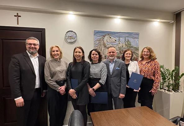 Siedem osób stoi w rzędzie w jasnym pokoju z jasnymi ścianami. Po lewej stronie wisi krzyż nad ciemnobrązowymi drzwiami, obok na ścianie znajduje się zegar. Na środku ściany wisi obraz przedstawiający białego konia i pejzaż. Trzy kobiety i dwóch mężczyzn trzymają teczki w rękach. Po prawej stronie stoi donica z zielonymi roślinami. Przed nimi znajduje się drewniany stół.