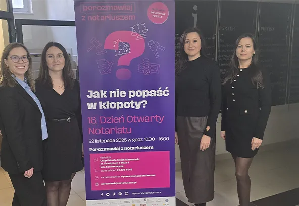 Cztery kobiety stoją obok siebie wewnątrz budynku przy stojaku z pionowym banerem o fioletowym tle i różowych elementach, promującym Dzień Otwarty Notariatu. Wszystkie mają na sobie ciemne stroje.