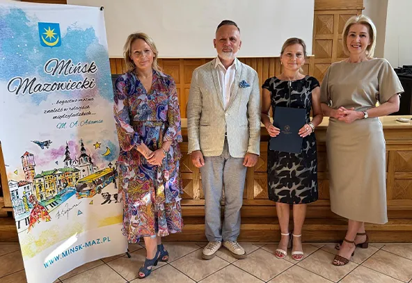 Cztery osoby stoją na tle drewnianej ściany i plakatu z napisem Mińsk Mazowiecki, w pomieszczeniu wewnętrznym.