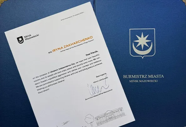 List od burmistrza Mińska Mazowieckiego skierowany do Iryny Zakharchenko z życzeniami i podpisem, na tle niebieskiej okładki z herbem miasta.
