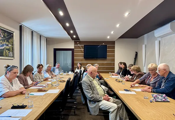 W sali konferencyjnej siedzi grupa osób przy długim stole, niektórzy piszą notatki, w tle znajduje się telewizor i okna z zasłonami.