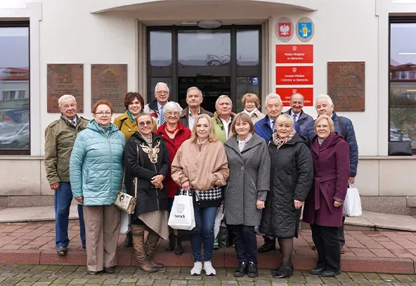 Na zewnątrz, w dzień grupa szesnastu osób stoi przed wejściem do budynku urzędu miasta. Większość osób ubrana w kurtki i płaszcze. W tle widoczne tablice z nazwami urzędu i herbem miasta.