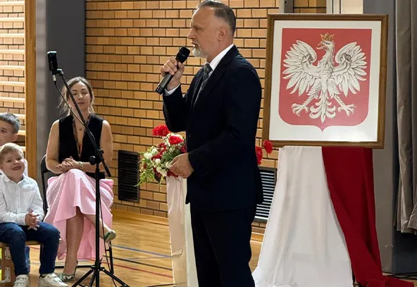 Mężczyzna w garniturze przemawia do mikrofonu w sali gimnastycznej, w tle kobieta i dziecko oraz godło Polski na stojaku ozdobione biało-czerwonym materiałem.